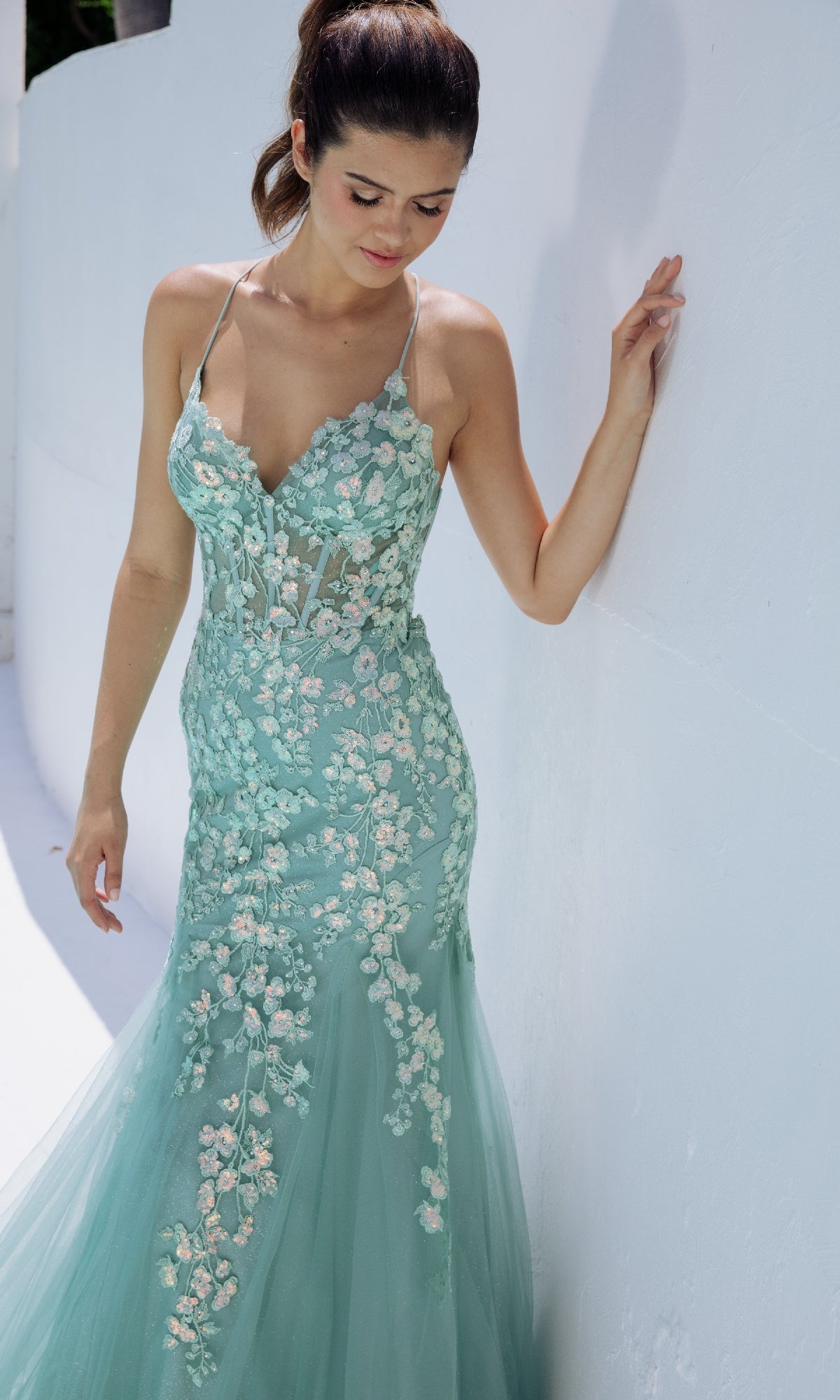 A woman wearing the Sequin-Floral Long Mermaid Prom Dress: Eureka 9696, a light teal gown with strappy back and floral embroidery, stands against a white wall, looking down.